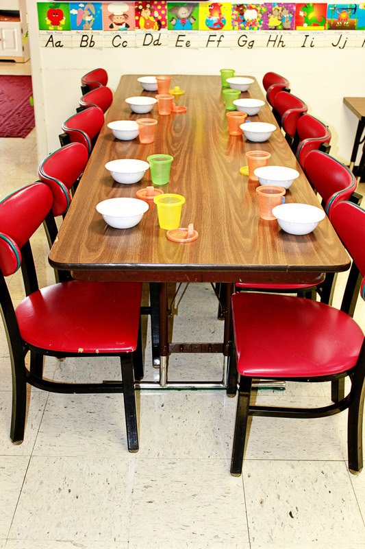 A classroom table set with bowls and colorful cups, surrounded by red chairs